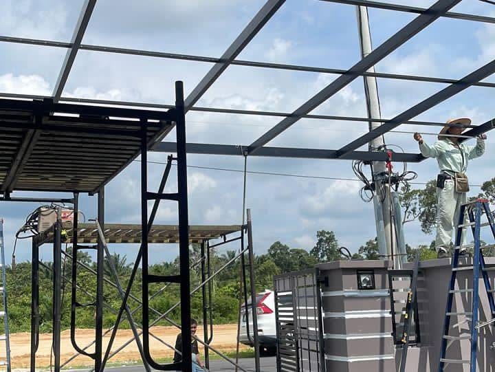 The welded dark steel roof frame of the porch part-built, with the first ceiling panels laid, mobile scaffolding and a worker on a step ladder at the frame.