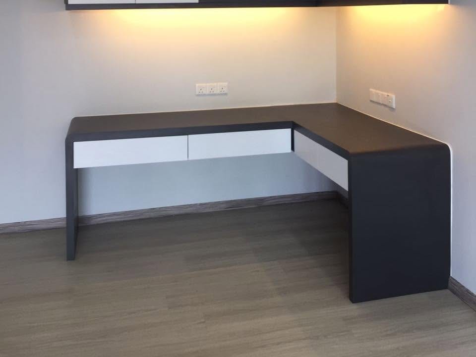 A built-in L-shaped desk with a dark grey wrap-around top, white drawer fronts and a warm-lit overhead unit, in an empty room with grey timber-look flooring.