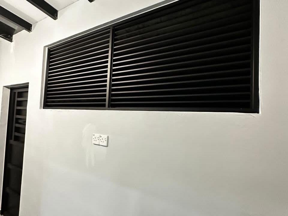 A wide two-panel dark aluminium louvre window high in a white wall, exposed dark ceiling beams above.