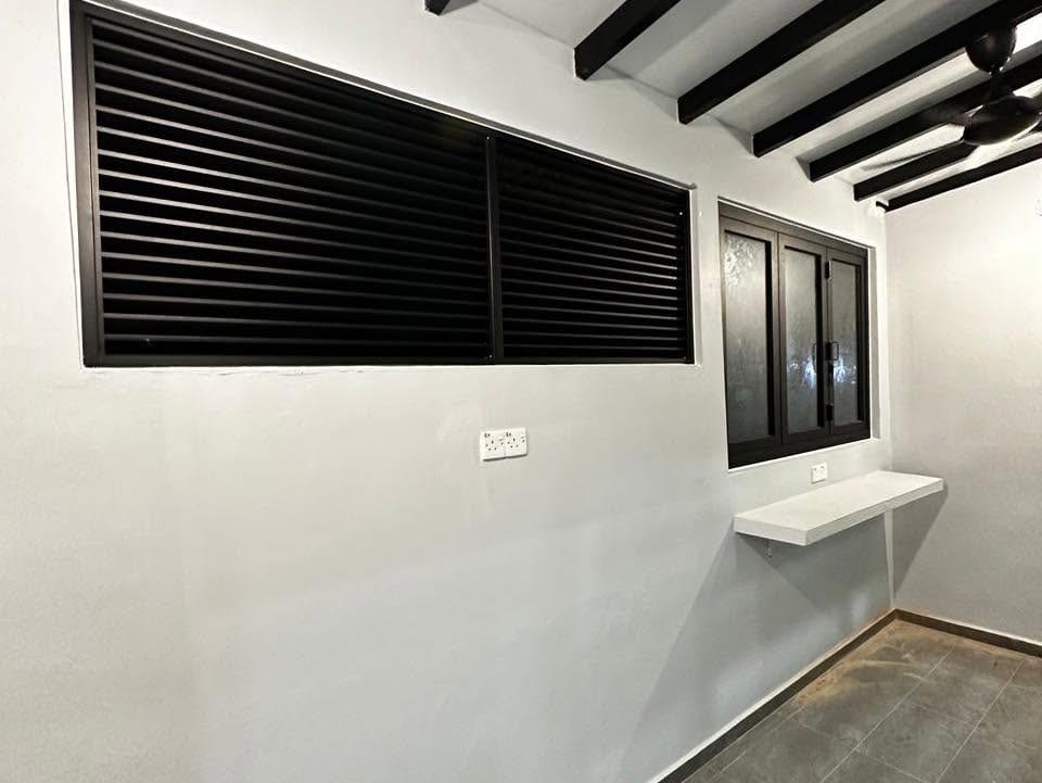 A yard corner with a large dark aluminium louvre window and a smaller casement window with obscured glass, a white wall shelf and a ceiling fan.